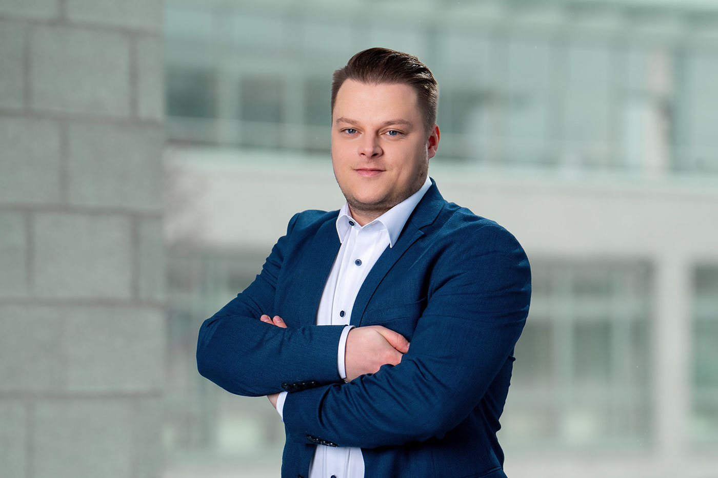 Porträt eines Mannes in Businesskleidung vor unscharfem Bürohintergrund. Er trägt ein dunkelblaues Sakko über einem weißen Hemd und steht mit verschränkten Armen, leicht lächelnd in selbstbewusster Haltung.