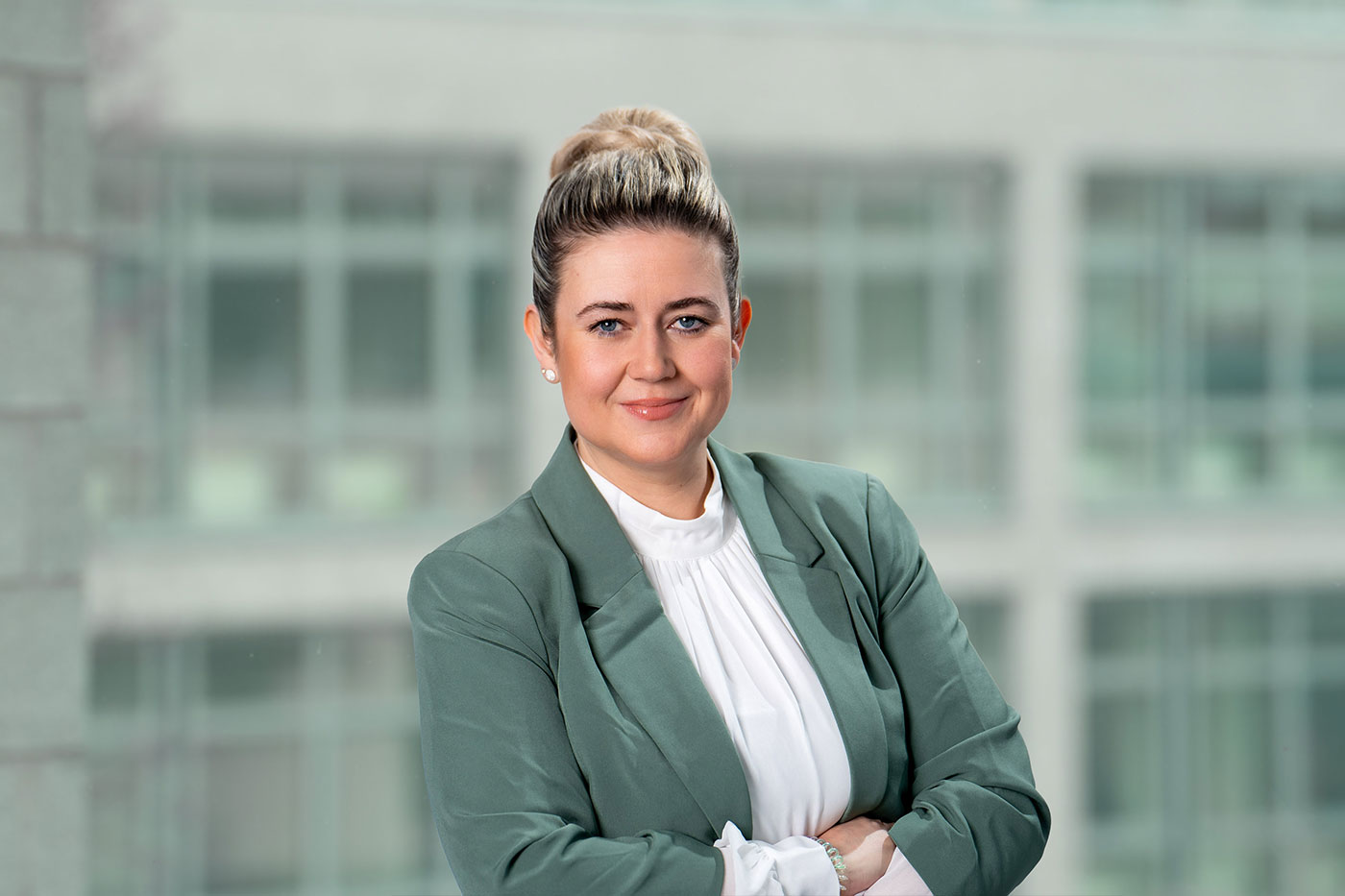Porträt einer Frau in Businesskleidung vor einem modernen, unscharfen Bürohintergrund. Sie trägt einen hellgrünen Blazer über einer weißen Bluse mit Stehkragen und blickt selbstbewusst mit einem freundlichen Lächeln in die Kamera.