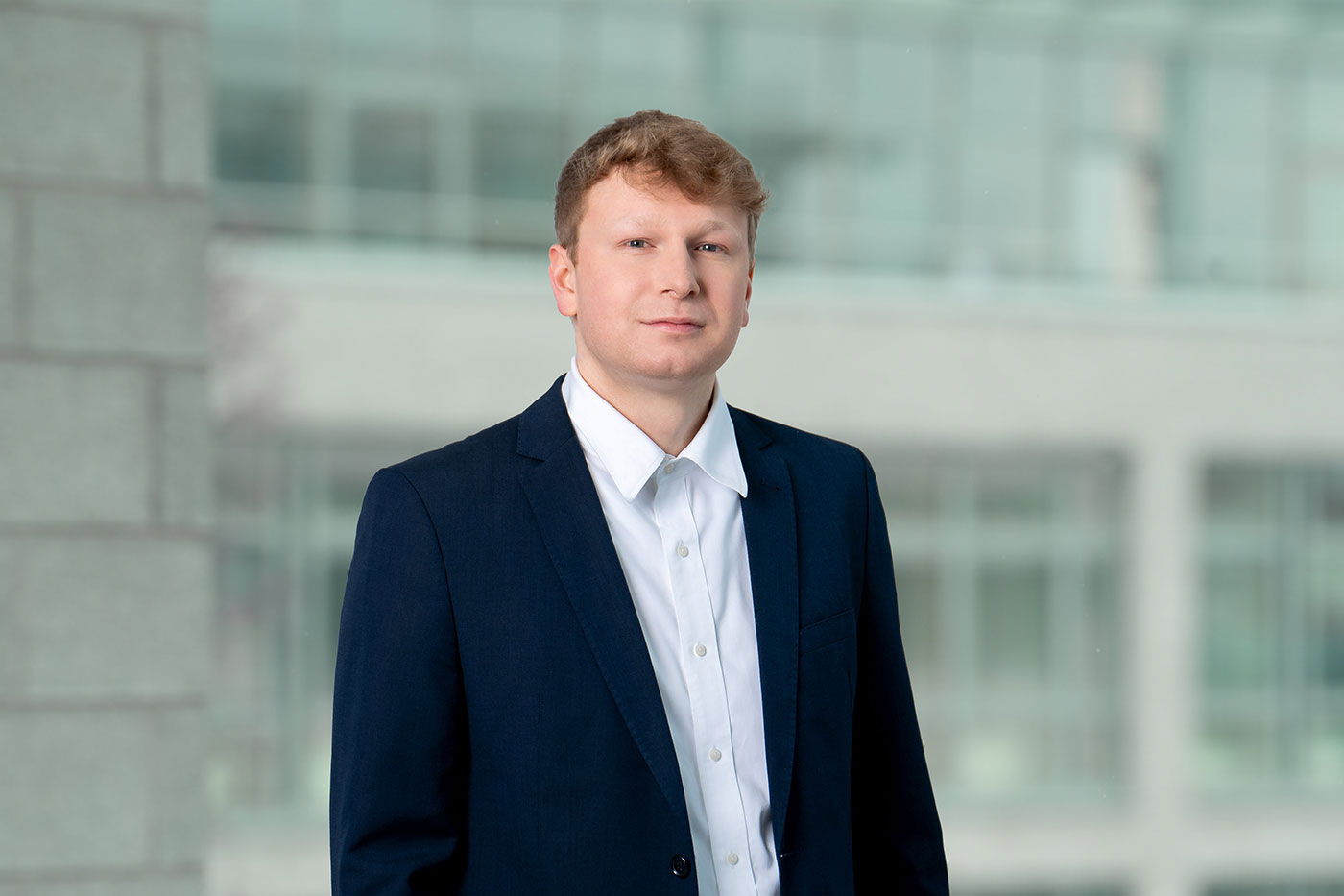 Porträt eines Mannes in Businesskleidung vor einem unscharfen, modernen Bürohintergrund. Er trägt ein dunkelblaues Sakko über einem weißen Hemd und blickt ruhig und professionell in die Kamera.