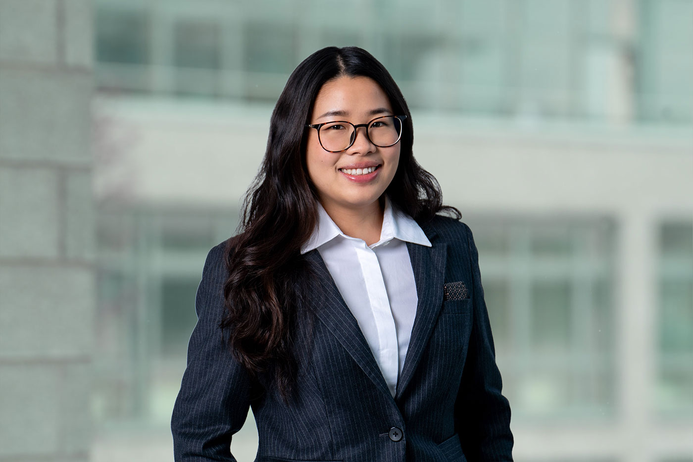 Porträt einer Frau in Businesskleidung vor unscharfem Bürohintergrund. Sie trägt einen dunklen Nadelstreifenblazer, ein weißes Hemd und eine Brille. Sie lächelt freundlich in die Kamera, ihre langen dunklen Haare fallen locker über die Schultern.