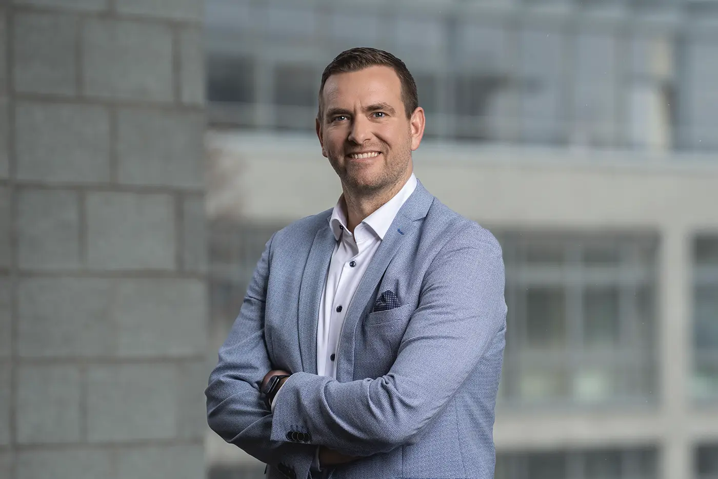 Ein Mann steht lächelnd mit verschränkten Armen vor einem unscharfen Hintergrund aus Glas- und Betonarchitektur. Er trägt ein hellblaues Sakko, ein weißes Hemd und ein Einstecktuch in der Brusttasche.