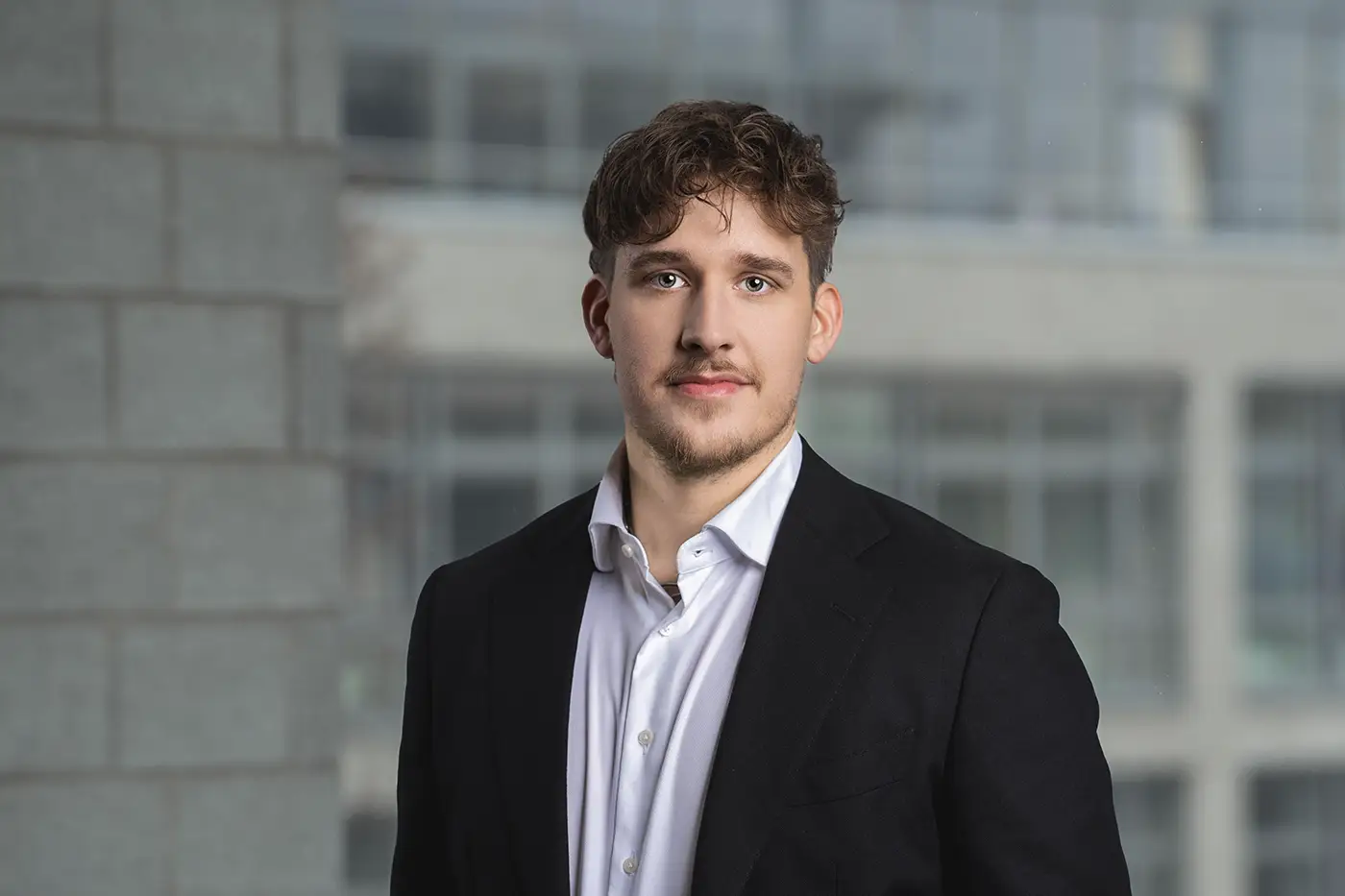 Ein Mann mit kurzen, lockigen Haaren steht in Businesskleidung vor einem unscharfen Bürogebäude-Hintergrund. Er trägt ein schwarzes Sakko über einem weißen Hemd und blickt ruhig in die Kamera.
