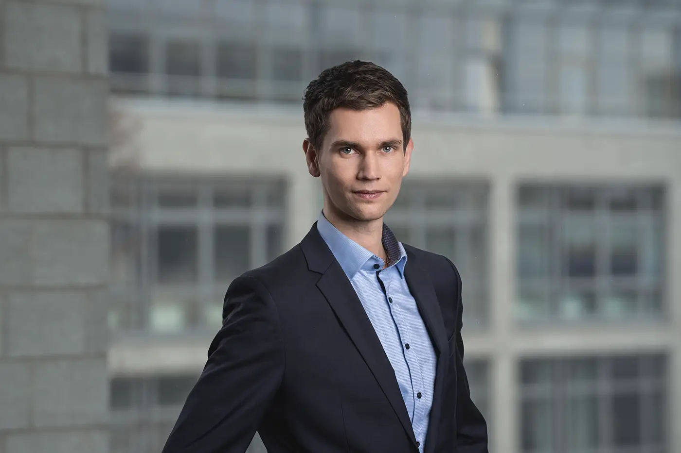 Ein Mann steht in Businesskleidung vor einem unscharfen Bürogebäude-Hintergrund. Er trägt ein dunkelblaues Sakko über einem hellblauen Hemd und blickt direkt in die Kamera mit leichtem Lächeln.