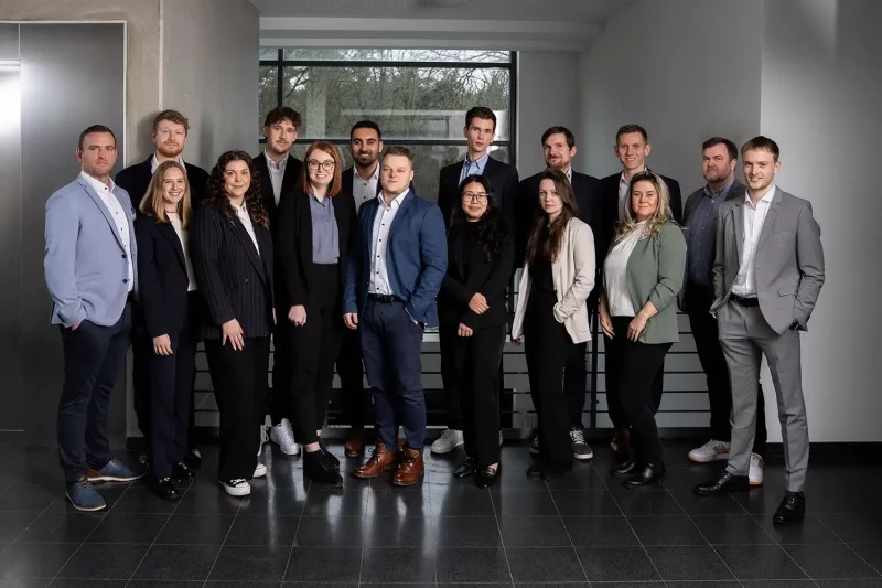 Gruppenfoto von 14 Personen in Businesskleidung, die in einem modernen Büroflur stehen. Die Gruppe ist gemischt, trägt überwiegend Anzüge, Blazer und Hemden in Blau-, Grau- und Beigetönen und blickt geschlossen in die Kamera. Im Hintergrund sind Fenster mit Blick auf eine Baumlandschaft zu sehen.