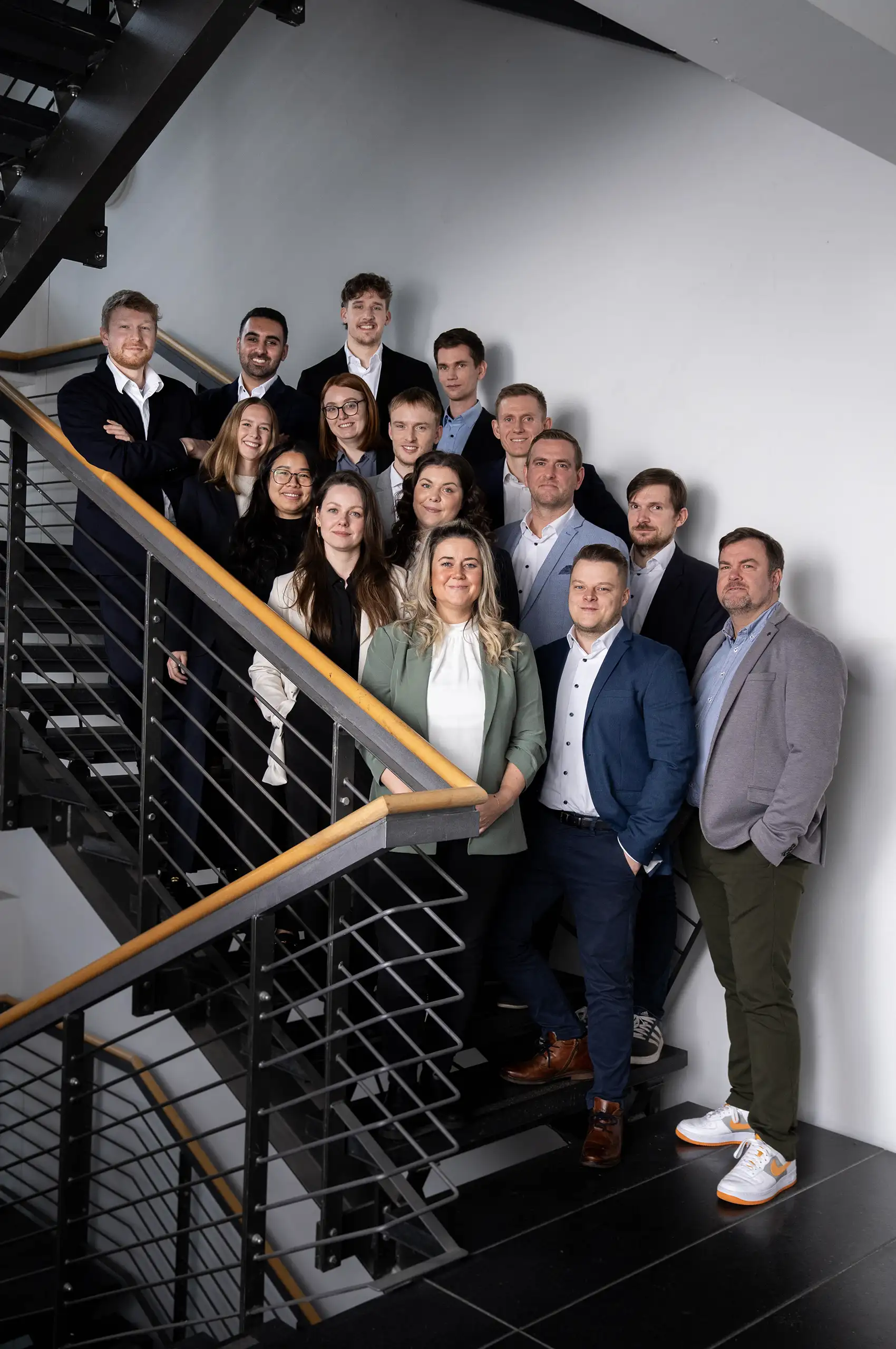 Gruppenfoto von 14 Personen in Businesskleidung, die gemeinsam auf einer modernen Treppe posieren. Die Gruppe steht gestaffelt auf den Stufen und blickt geschlossen in die Kamera. Die Personen tragen Anzüge, Blazer und Business-Outfits in gedeckten Farben. Der Hintergrund ist eine weiße Wand mit schwarzem Metallgeländer und Holzhandlauf.