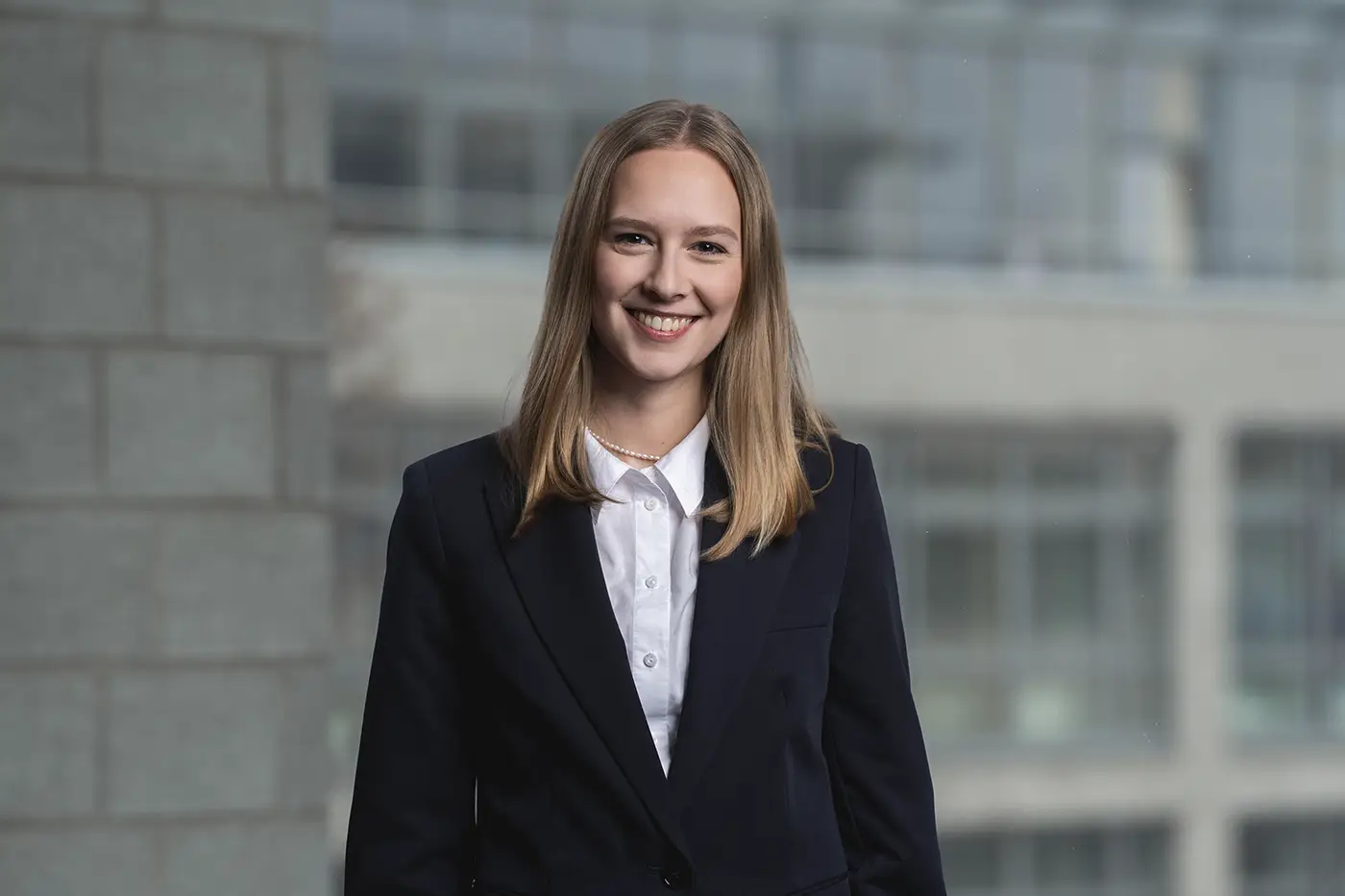 Eine Frau steht in Businesskleidung vor einem unscharfen Bürogebäude-Hintergrund. Sie trägt ein dunkles Sakko über einer weißen Bluse mit Perlenkette und lächelt freundlich in die Kamera.
