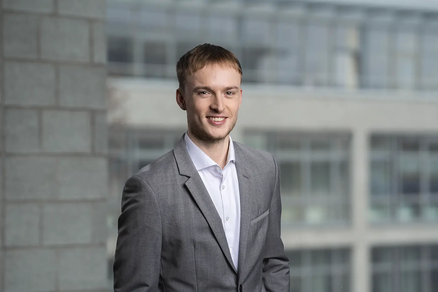 Ein Mann steht in einem grauen Anzug mit weißem Hemd vor einem unscharfen Hintergrund aus Glas- und Betonarchitektur. Er lächelt freundlich und blickt direkt in die Kamera.