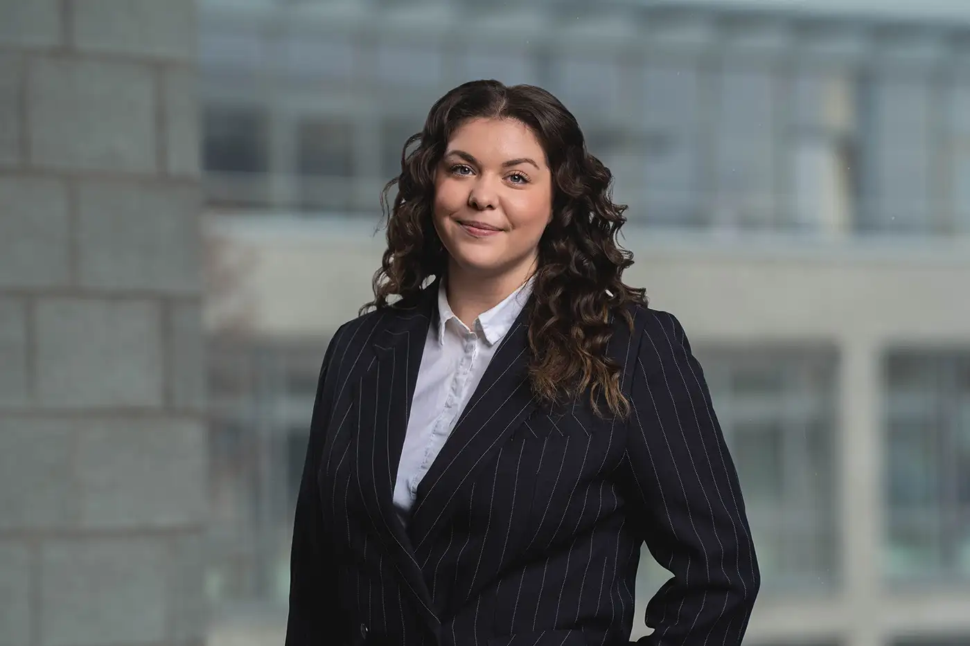 Eine Frau mit dunklen, lockigen Haaren steht vor einem unscharfen Hintergrund aus Glas- und Betonarchitektur. Sie trägt ein dunkelblaues, fein gestreiftes Business-Jackett über einer weißen Bluse und lächelt dezent in die Kamera.