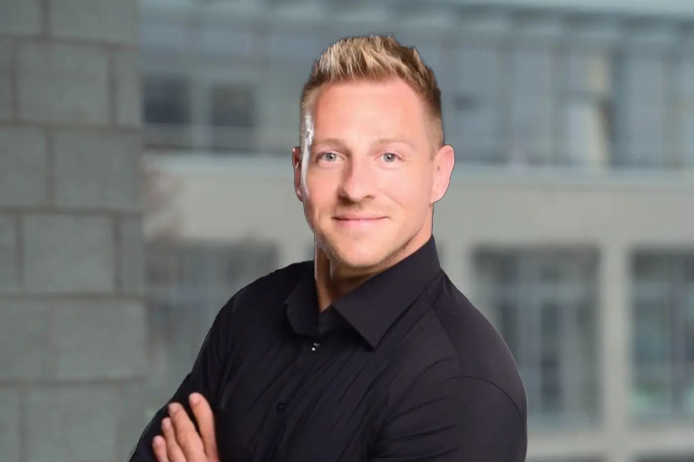 Ein Mann mit kurz geschnittenem, blondem Haar steht lächelnd vor einem unscharfen Hintergrund aus Glas- und Betonarchitektur. Er trägt ein schwarzes Hemd und hat die Arme vor dem Körper verschränkt.