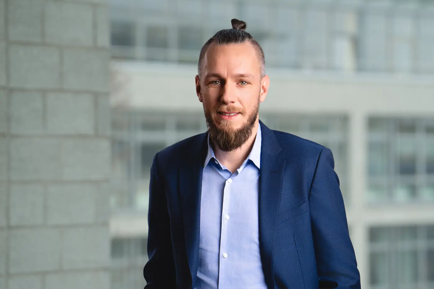 Porträt eines Mannes mit zusammengebundenem Oberkopf-Haar (Man-Bun) und Vollbart. Er trägt ein hellblaues Hemd und ein dunkelblaues Sakko. Der Hintergrund zeigt ein modernes Bürogebäude mit Glasfassade.