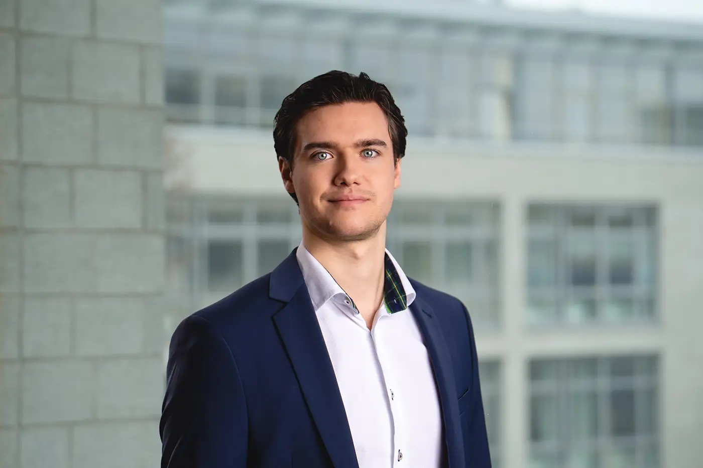Porträt eines jungen Mannes im Business-Outfit vor einem modernen Bürogebäude mit Glasfassade. Er trägt ein dunkelblaues Sakko und ein weißes Hemd, blickt freundlich und selbstbewusst in die Kamera.