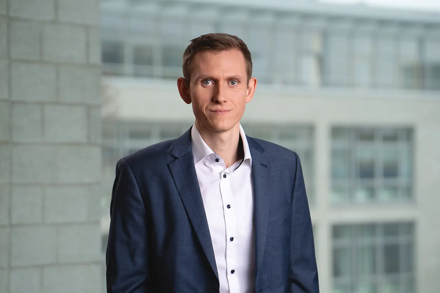 Porträt eines jungen Mannes mit kurzem braunem Haar, hellem Teint und neutralem Gesichtsausdruck. Er trägt ein weißes Hemd mit dunklen Knöpfen und ein dunkelblaues Sakko. Der Hintergrund zeigt ein modernes Bürogebäude mit großen Fenstern.