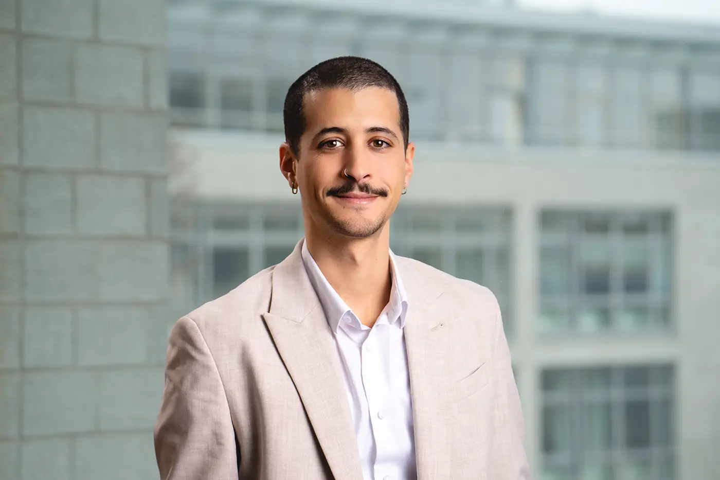 Porträt eines jungen Mannes mit kurz rasiertem Haar, Ohrringen und Nasenring. Er trägt ein beiges Sakko und ein weißes Hemd. Im Hintergrund ist ein modernes Bürogebäude mit großen Fenstern zu sehen.
