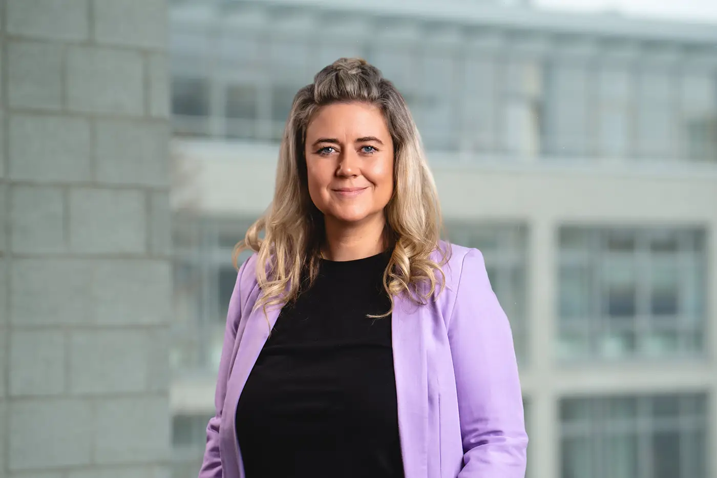 Porträt einer lächelnden Frau mit langen, gewellten blonden Haaren. Sie trägt ein schwarzes Oberteil und ein fliederfarbenes Blazerjackett. Der Hintergrund zeigt ein modernes Bürogebäude mit großen Glasfenstern.