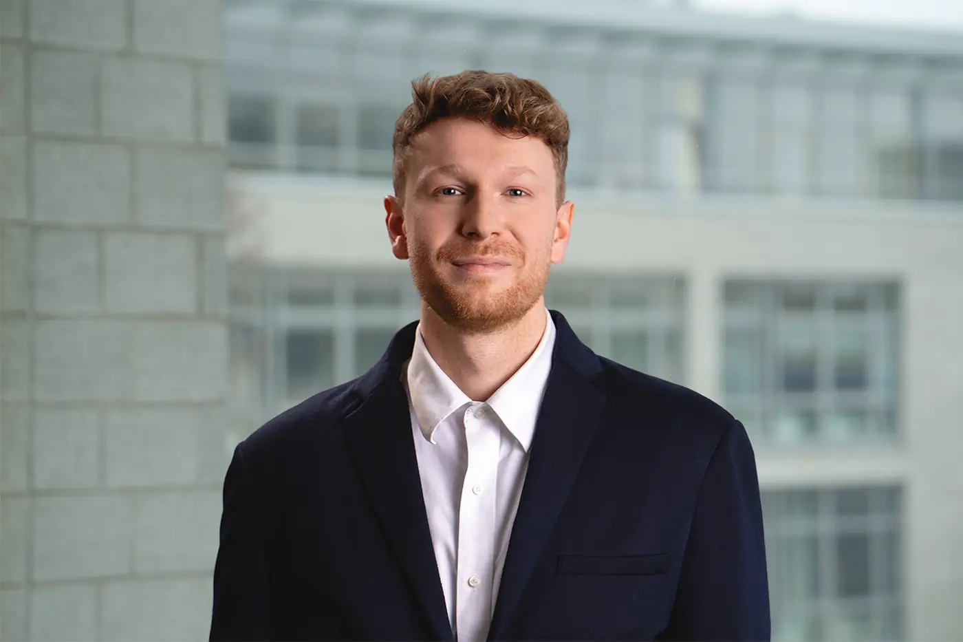 Porträt eines jungen Mannes mit rötlich-blondem Haar, Bart und einem selbstsicheren Lächeln. Er trägt ein dunkelblaues Sakko und ein weißes Hemd, im Hintergrund ein modernes Bürogebäude mit Glasfassade.