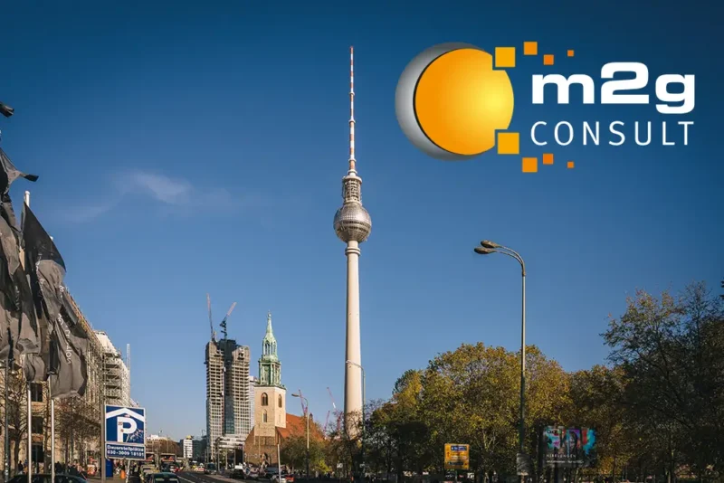 Blick auf den Berliner Fernsehturm bei klarem Himmel, rechts oben eingeblendet das Logo von „m2g CONSULT“ mit stilisierter oranger Kugel und digitalen Quadraten. Im Vordergrund Straßenszene mit Gebäuden, Autos und Bäumen.