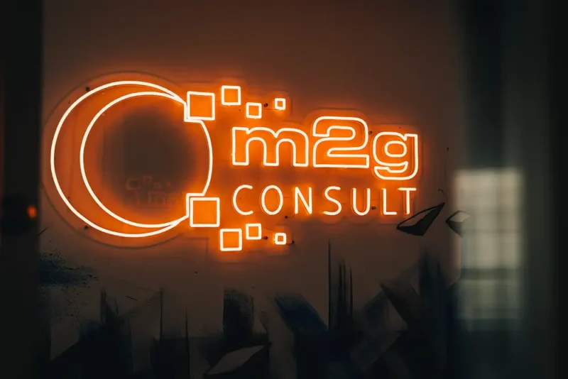 Leuchtendes Neon-Logo an einer Wand mit der Aufschrift „m2g CONSULT“ in orangefarbenem Licht. Die Gestaltung enthält digitale Quadrate, die aus einem großen offenen Kreis auf der linken Seite hervorgehen. Der Hintergrund ist dunkel mit angedeuteter urbaner Struktur.