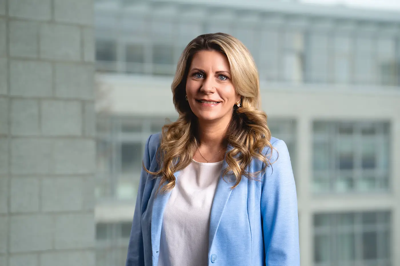 Porträt einer Frau mit langen, gewellten, blonden Haaren. Sie trägt ein hellblaues Jackett über einer weißen Bluse. Der Hintergrund zeigt ein modernes Bürogebäude mit großen Fenstern.