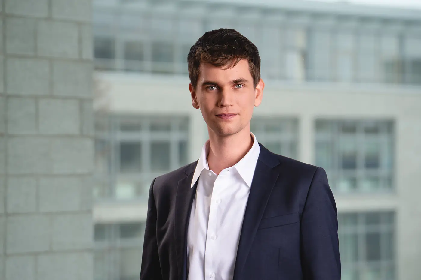 Porträt eines jungen Mannes mit kurzen braunen Haaren und hellem Teint, der ruhig in die Kamera blickt. Er trägt ein dunkelblaues Sakko und ein weißes Hemd, im Hintergrund ein modernes Bürogebäude mit großen Fenstern.