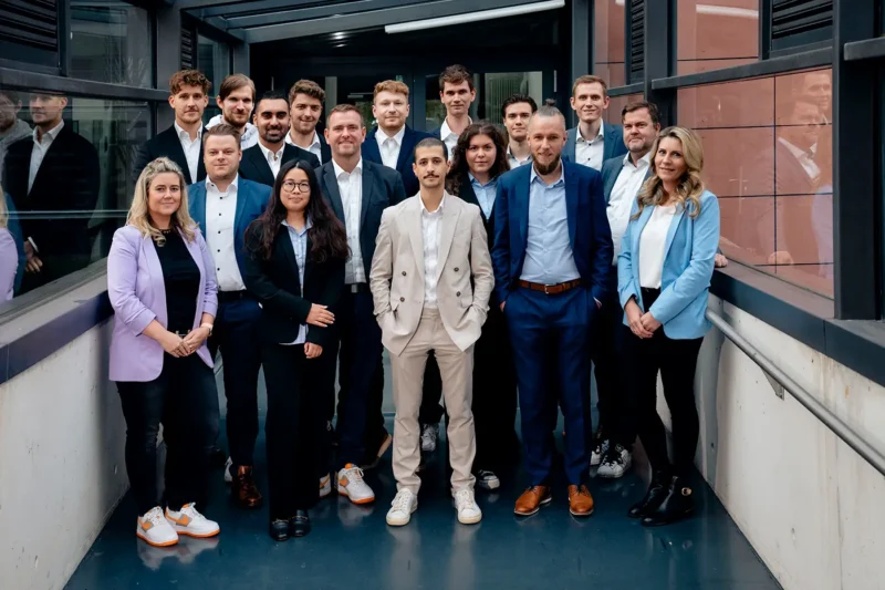 Gruppenfoto eines etwa 18-köpfigen, gemischten Teams in Businesskleidung. Die Personen stehen in einem modernen Bürogebäude im Glasflur. Die Kleidung reicht von Anzügen bis hin zu sportlich-elegantem Stil. Im Zentrum steht eine Person in einem beigen Anzug, umgeben von Kolleginnen und Kollegen.
